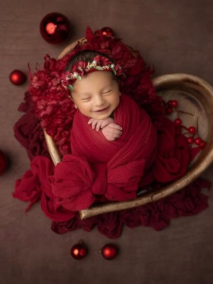 A beautiful smile from a newborn nestled in a heart shaped bowl, surrounded by festive red ornaments. This is the perfect way to celebrate your baby's first holiday season.