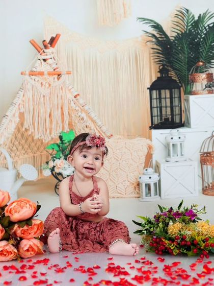 A joyful baby girl sitting on the floor amidst rose petals in a boho-themed setup. Her happy expression and the beautiful decor create a heartwarming image.