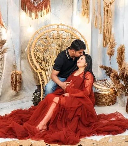 A tender moment in a boho-chic studio setting. The partner kisses the expecting mother's forehead as she relaxes in a large wicker chair, draped in a beautiful red gown.