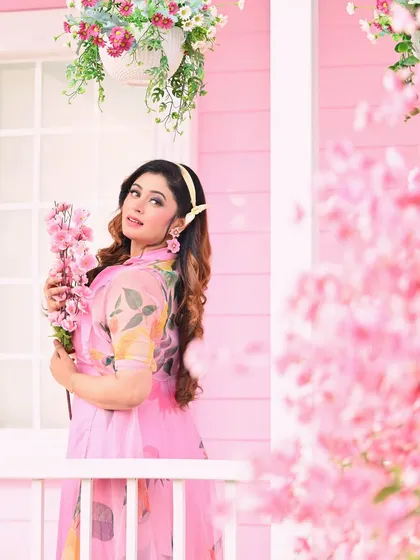 A whimsical fashion portrait set against a dreamy pink cherry blossom backdrop. The model's floral dress and the pastel setting create a soft and romantic atmosphere.