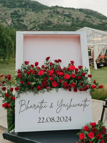 A simple yet powerful welcome sign for an outdoor wedding. The sign, featuring the couple's names, is framed with a beautiful arrangement of red roses, making a bold and romantic statement.