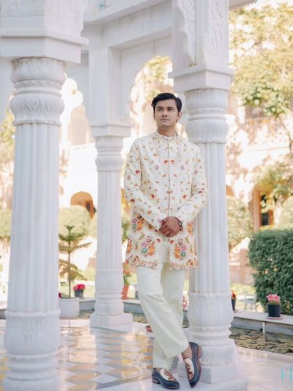 The groom stands poised and elegant in his embroidered sherwani, framed by the white marble pillars of a Rajasthani palace.
