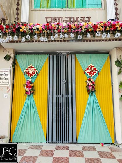 A traditional and colorful entrance decoration for a Haldi ceremony at home. The main gate is adorned with yellow and turquoise drapes and floral accents, welcoming guests with festive cheer.