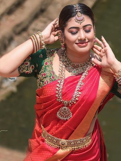 A traditional portrait of a model in a red and green saree, taken outdoors near a rustic wall. The focus is on the rich colors and textures of the ethnic wear.