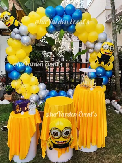 A fun Minions-themed party setup for an outdoor space. The decor uses the signature yellow and blue colors, along with Minion foil balloons and themed table covers.