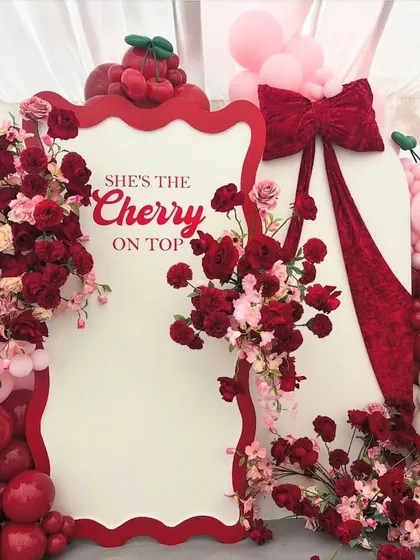 "She's the Cherry on Top!" This adorable and unique backdrop features a custom-shaped frame, red and pink balloons, and beautiful floral arrangements with cherry details.