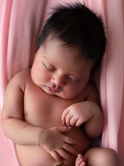 A close-up of a three-week-old baby girl, sleeping peacefully in a pink sling. The focus is on her serene expression and the soft, womb-like environment.