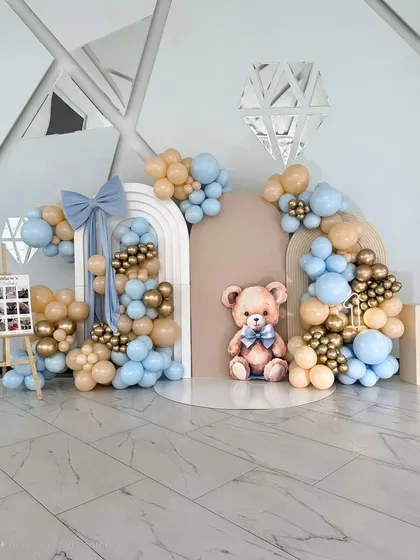 A sweet and classic teddy bear theme for a first birthday. The backdrop features arched panels, a large teddy bear cutout, and a beautiful balloon garland in pastel blue, beige, and gold.