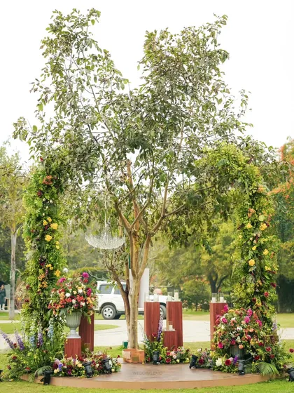 A beautiful mandap setup under a natural tree arch. The space is defined by lush floral pillars, creating an intimate and organic setting for the wedding vows.