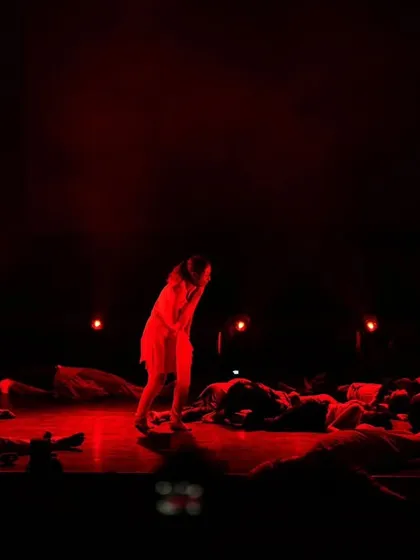 Bathed in red light, a solo performer stands amidst a group in a scene depicting struggle and resilience. This highlights our use of lighting and stage composition to evoke strong emotions and tell a compelling story through dance.