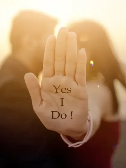 A creative and symbolic shot perfect for an engagement or pre-wedding announcement. The focus is on her hand with "Yes I Do!" written on it, with the couple blurred softly in the background against a sunset.