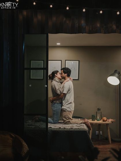 A romantic kiss in a softly lit bedroom. The framing through the doorway creates a sense of looking in on a private, intimate moment.