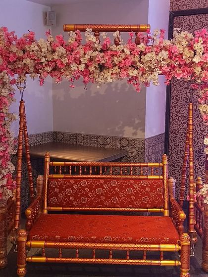A beautifully decorated traditional wooden jhula (swing) for a baby shower ceremony. The swing is adorned with an arch of pink and white cherry blossoms.
