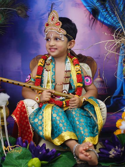 A thoughtful pose from the little boy dressed as Krishna. The detailed setup with brass lamps and lotus flowers adds to the authenticity of the theme.
