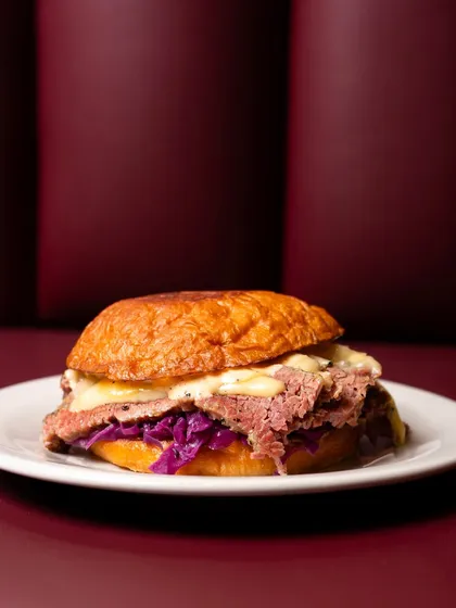 This pastrami sandwich is shot against a rich, deep red background that complements the colors in the food. The lighting highlights the melted cheese and juicy meat.