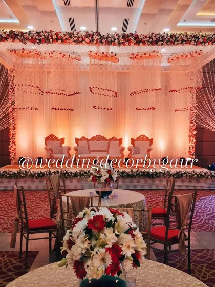 A classic indoor wedding stage setup with a red and white floral mandap, showing the guest seating and table centerpieces in the foreground.