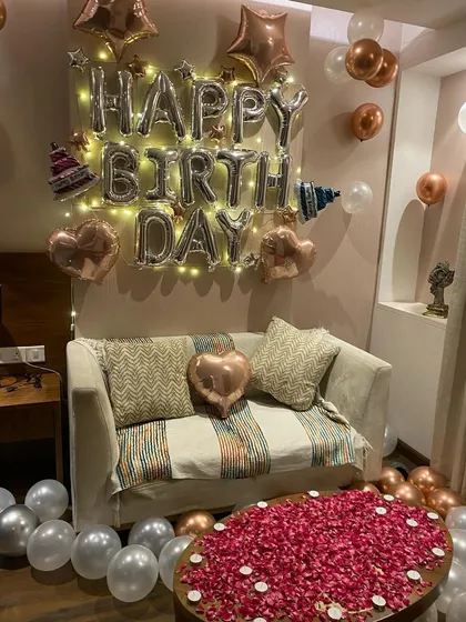 The celebration corner of the room. A cozy sofa is framed by "Happy Birthday" foil balloons with integrated fairy lights, while the coffee table is covered in rose petals, creating a perfect spot for cake cutting and photos.