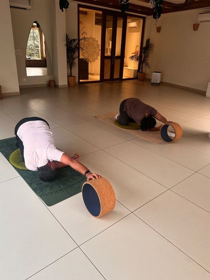 Two students use yoga wheels to deepen their child's pose, opening up the shoulders and upper back.