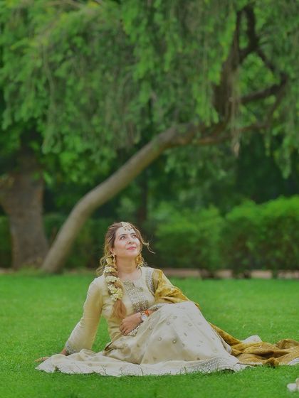 A dreamy, cinematic shot of a woman in a beautiful lehenga, sitting on the grass in a park. Her upward glance and the soft green background create a fairytale-like atmosphere.