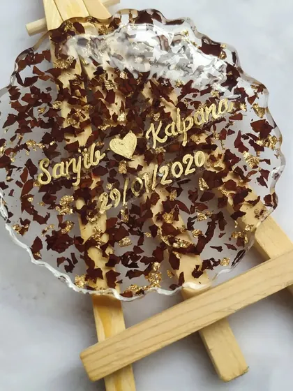 A close-up of a single coaster made from preserved flower petals and gold leaf. Personalized with the couple's names, it serves as a small, beautiful reminder of their special day.