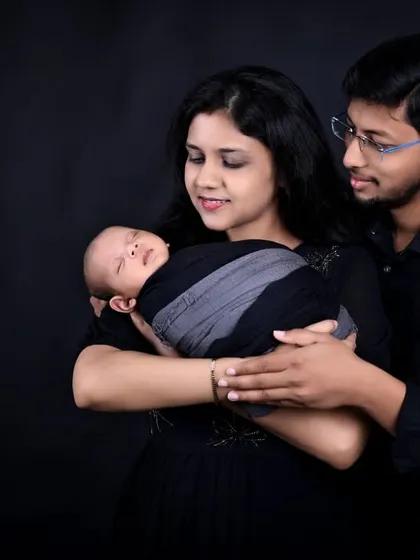 A beautiful composition of a new family. The mother holds their sleeping newborn while the father stands behind, looking over them with a gentle smile, all set against a classic dark backdrop.