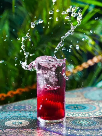 A dynamic splash frozen in time in a glass of red cocktail. The motion of the liquid creates an exciting and refreshing visual against the tropical green background, perfect for an energetic ad campaign.