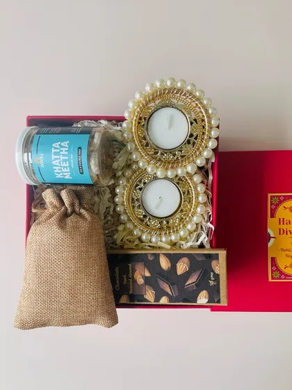 A compact and beautiful Diwali hamper in a red box. It includes pearl T-light holders, a chocolate bar, a snack mix, and a potli of nuts.