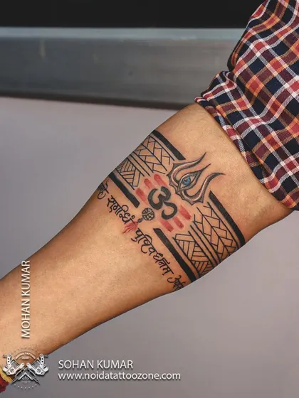 This is a custom band tattoo with a Lord Shiva theme. It incorporates a Trishul, the Om symbol, and the Mahamrityunjaya mantra into a single, cohesive armband design with red and black ink.