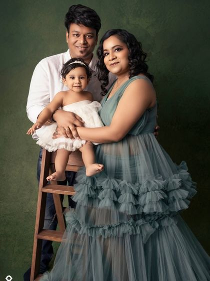 A family of three posing together against a rich green backdrop. The mother's elegant ruffled gown complements the classic look of her husband and daughter.