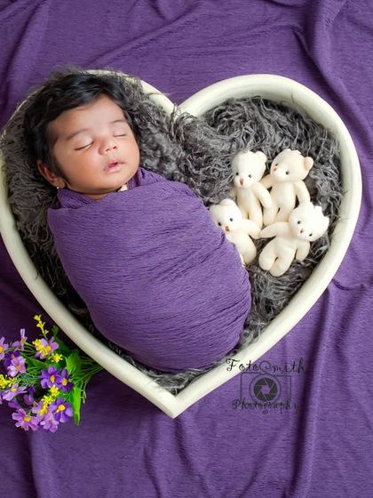 A baby swaddled in purple, nestled in a heart-shaped bowl with tiny teddy bears. The rich purple background makes this a vibrant and loving portrait.