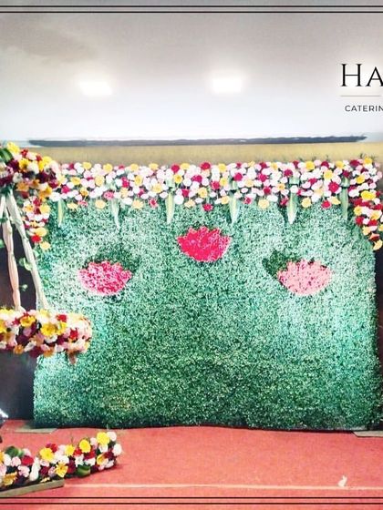 A naming ceremony stage featuring a multi-colored floral moon cradle. The green backdrop is decorated with lotus flower cutouts, adding a traditional and symbolic touch.