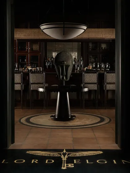 The dramatic entrance to the bar at Lord Elgin, framed to create a powerful first impression. A sculptural artifact stands at the center, drawing the eye towards the classic, wood-paneled bar in the background.
