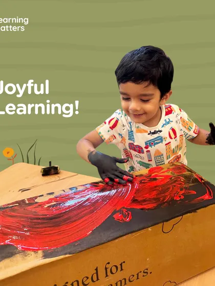 With hands protected by gloves, this child joyfully paints a cardboard box. This engagement is not about creating a masterpiece, but about the sensory experience of spreading paint and the freedom to transform an everyday object into something new.