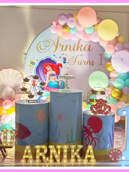 A pastel-colored mermaid-themed first birthday. The setup features an Ariel backdrop, an octopus balloon sculpture, and a large light-up number '1'.