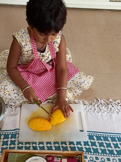 A child carefully practices her knife skills, slicing mangoes for our kitchen activity. Using child-safe tools, I guide them in developing practical life skills, confidence, and fine motor control in a supervised environment.