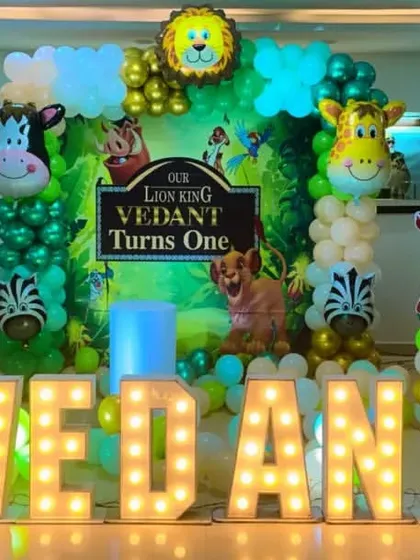 A Lion King first birthday party theme. This grand setup includes large marquee letters spelling the name 'VEDANT' and balloon columns with animal character foils.