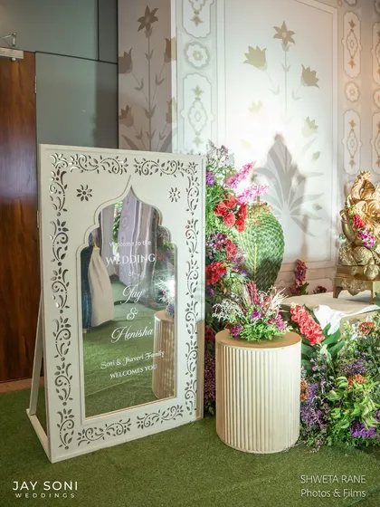 Personal touches make an entrance special. For this wedding, I placed a beautiful welcome mirror with intricate jaali-work details amidst a garden of colorful flowers, offering a warm and personalized greeting.