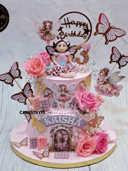 An elegant two-tier butterscotch cake for a third birthday. It's topped with a 3D girl figurine and decorated with fresh roses, baby's breath, and delicate paper fairies and butterflies.