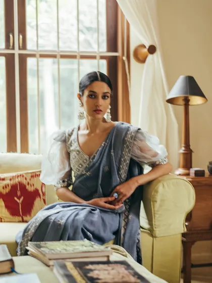 An elegant indoor portrait from a lookbook. The model is seated in a well-lit living room, showcasing a modern saree with puffed sleeves in a sophisticated, natural setting.