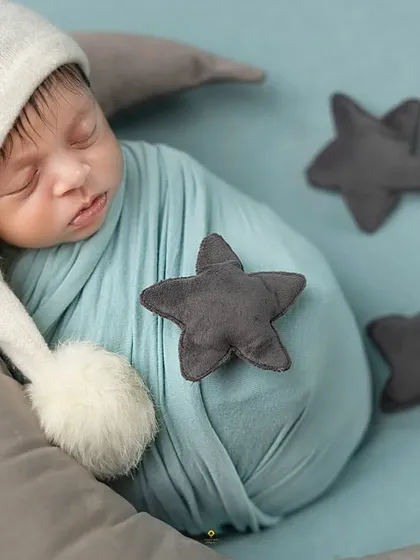 Dreaming among the stars. This baby is swaddled in blue and wearing a sleepy cap, nestled on a moon prop with soft, grey stars scattered around.