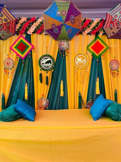 Here is a classic Haldi and Mehndi seating arrangement. The backdrop combines bright yellow and green drapes with traditional Rajasthani umbrellas, and I provide a covered chowki or sofa for the main ceremony, complete with matching cushions.