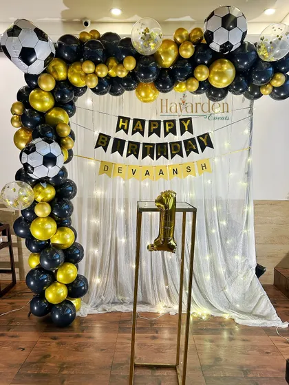 A football-themed party for a first birthday. The black and gold balloon arch is decorated with football balloons, set against a curtain with fairy lights.