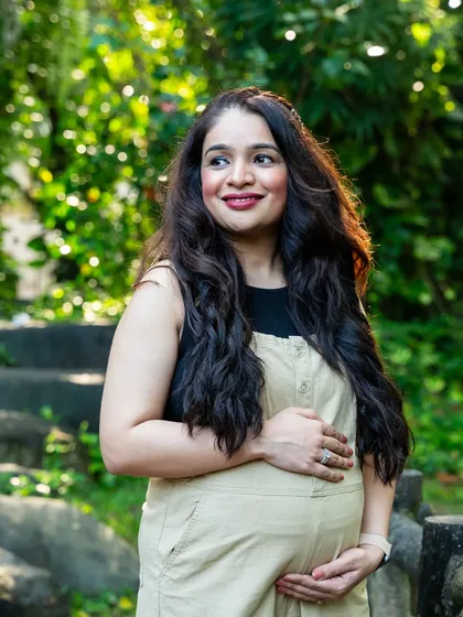 A candid moment of the mom-to-be, glowing with happiness in a casual beige jumpsuit, surrounded by the fresh greenery of a park.