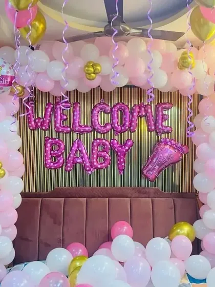 An elaborate "Welcome Baby" room decoration in pink and white. The setup features balloon pillars, foil letters against a gold fringe backdrop, and balloons on the ceiling.