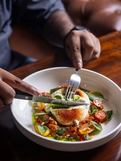 A refreshing bite of the Burrata Salad. A guest cuts into the creamy cheese, mixing it with the pesto and tomatoes.