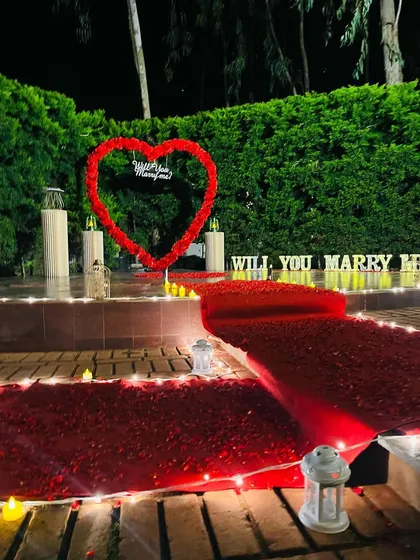 A different angle of the garden proposal setup, showing the detail of the red carpet aisle and the placement of lanterns and LED candles. The 'WILL YOU MARRY ME' marquee letters and floral heart are visible in the background, creating a layered and photogenic scene.