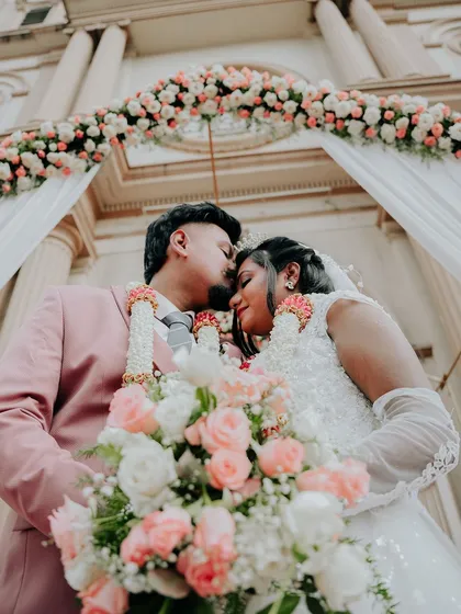 An intimate moment from a Christian wedding. The groom kisses the bride's forehead under a beautiful floral arch, with the grandeur of the church creating a timeless backdrop.