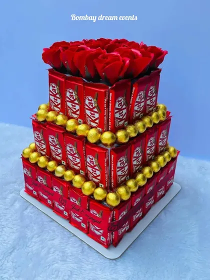 A classic three-tiered chocolate tower made with KitKats and Ferrero Rocher, topped with a bouquet of red roses. It's a perfect gift or centerpiece for any chocolate lover's celebration.
