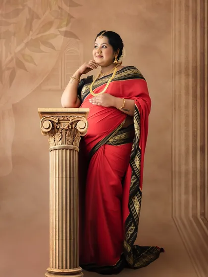 A thoughtful and elegant pose of a mom-to-be in a red saree, leaning against a classic pillar in a studio with a painted backdrop.