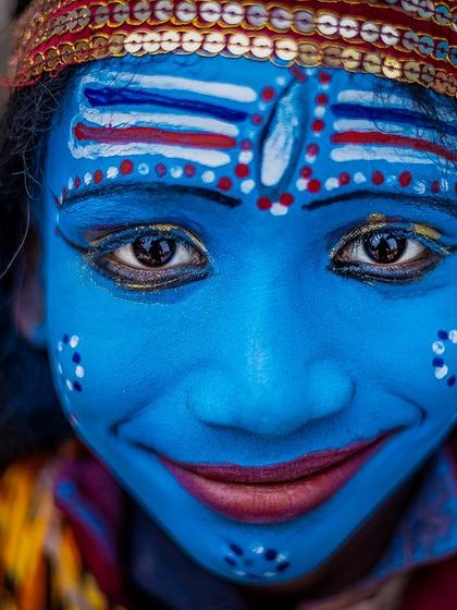 A child dressed as Lord Shiva smiles directly at the camera on the ghats of Banaras, his painted blue face and innocent eyes creating a delightful portrait.